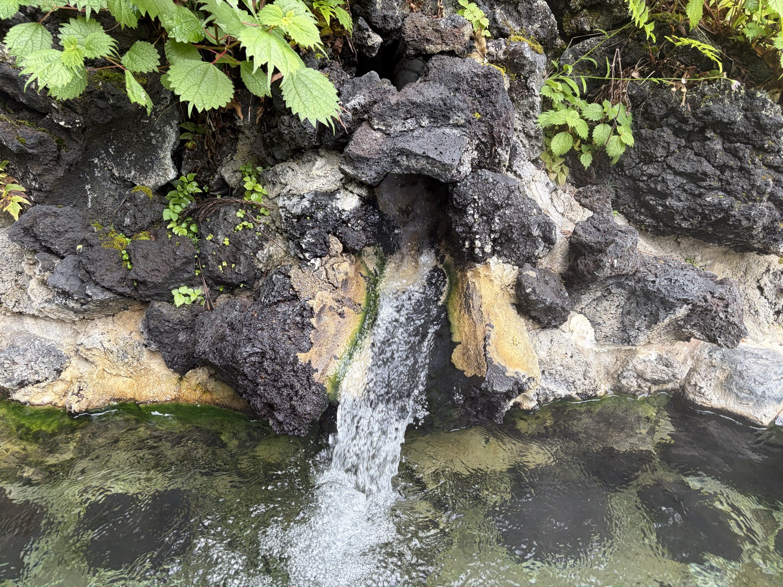 野天風呂 湯口アップ・黒い岩の隙間から湯が太い1本の流れで滝のように落ちる、白い析出物がうっすら付着