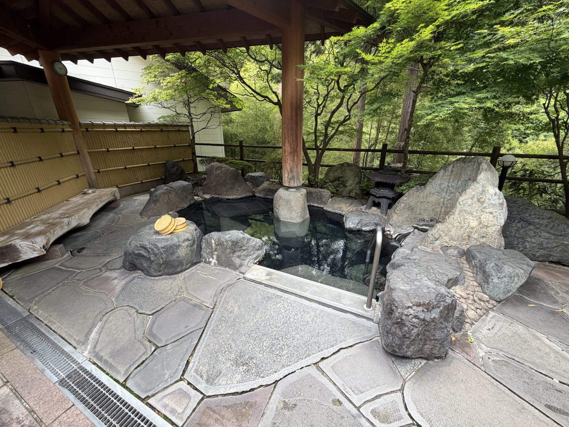 屋根付きの野天風呂。岩組みの湯船と竹垣、まわりはもみじの緑