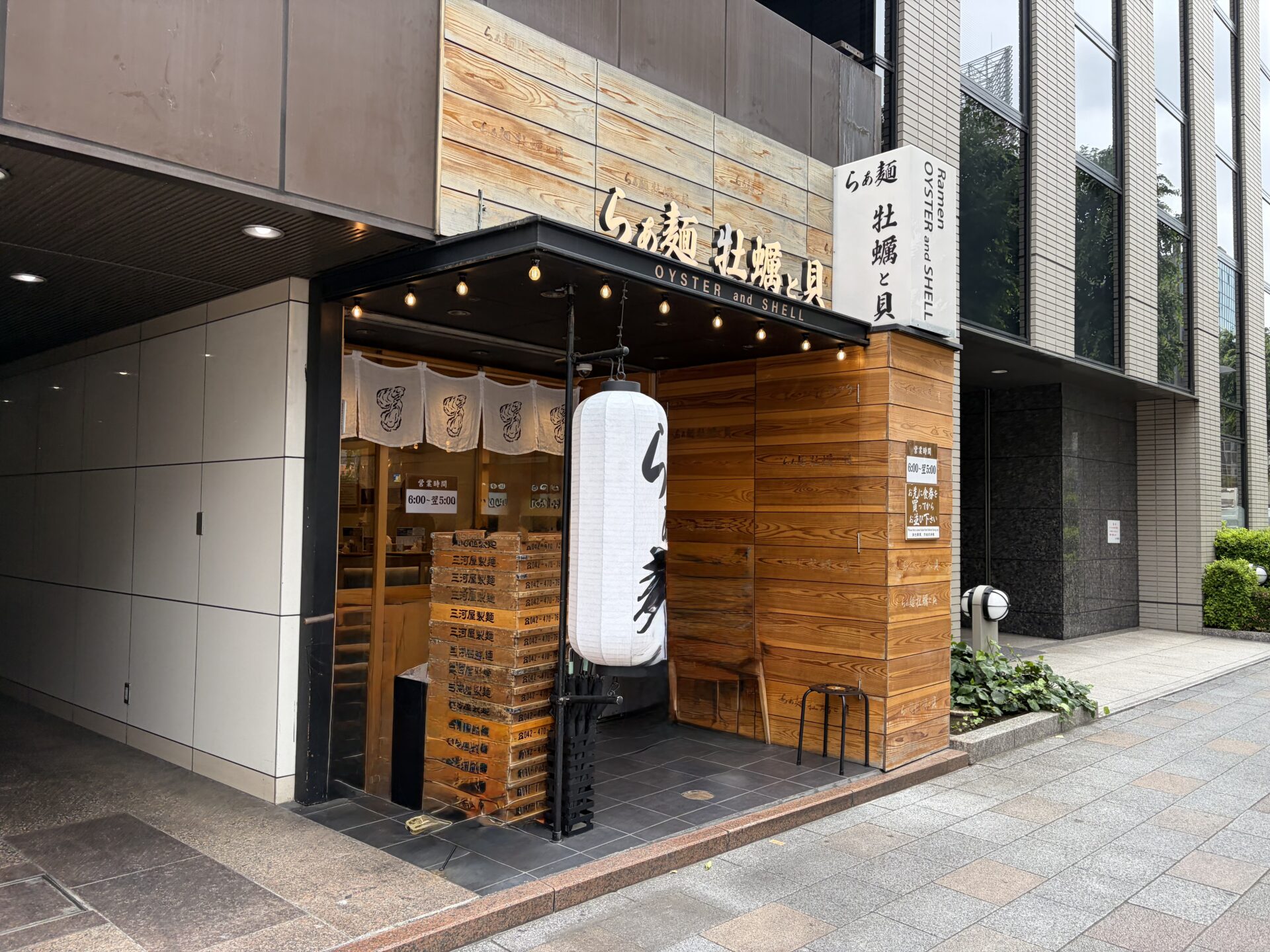 らぁ麺 牡蠣と貝の外観。白い大提灯と「らぁ麺 牡蠣と貝 OYSTER and SHELL」の看板、店先には三河屋製麺の麺箱、袖看板に営業時間6:00〜翌5:00
