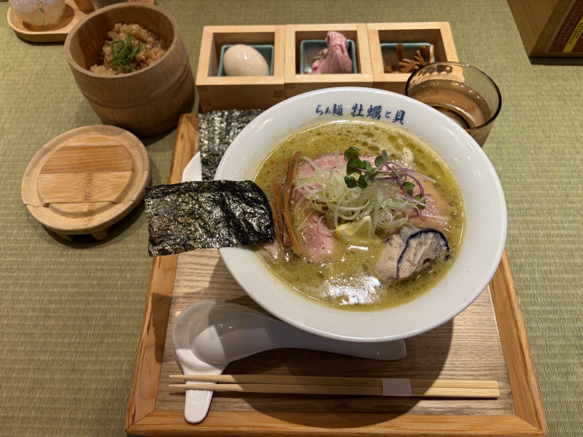 濃厚牡蠣らぁ麺の卓上一式。緑がかったスープにレモンスライス・レアめのチャーシュー・牡蠣・メンマ・白ねぎ・紫かいわれ、海苔は別添え。左奥に木桶入りの炊き込みご飯、木箱3連のトッピング(味玉・チャーシュー・メンマ)