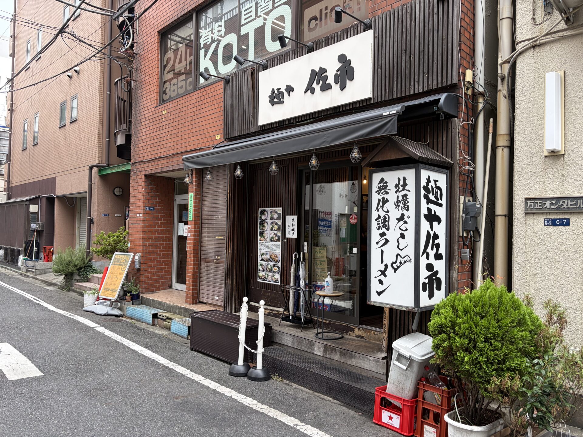 麺や佐市の外観。白地の店名看板と、袖看板の「牡蠣だし 無化調ラーメン」。入口には「生牡蠣あり〼」の貼り紙とENGLISH MENUのステッカー