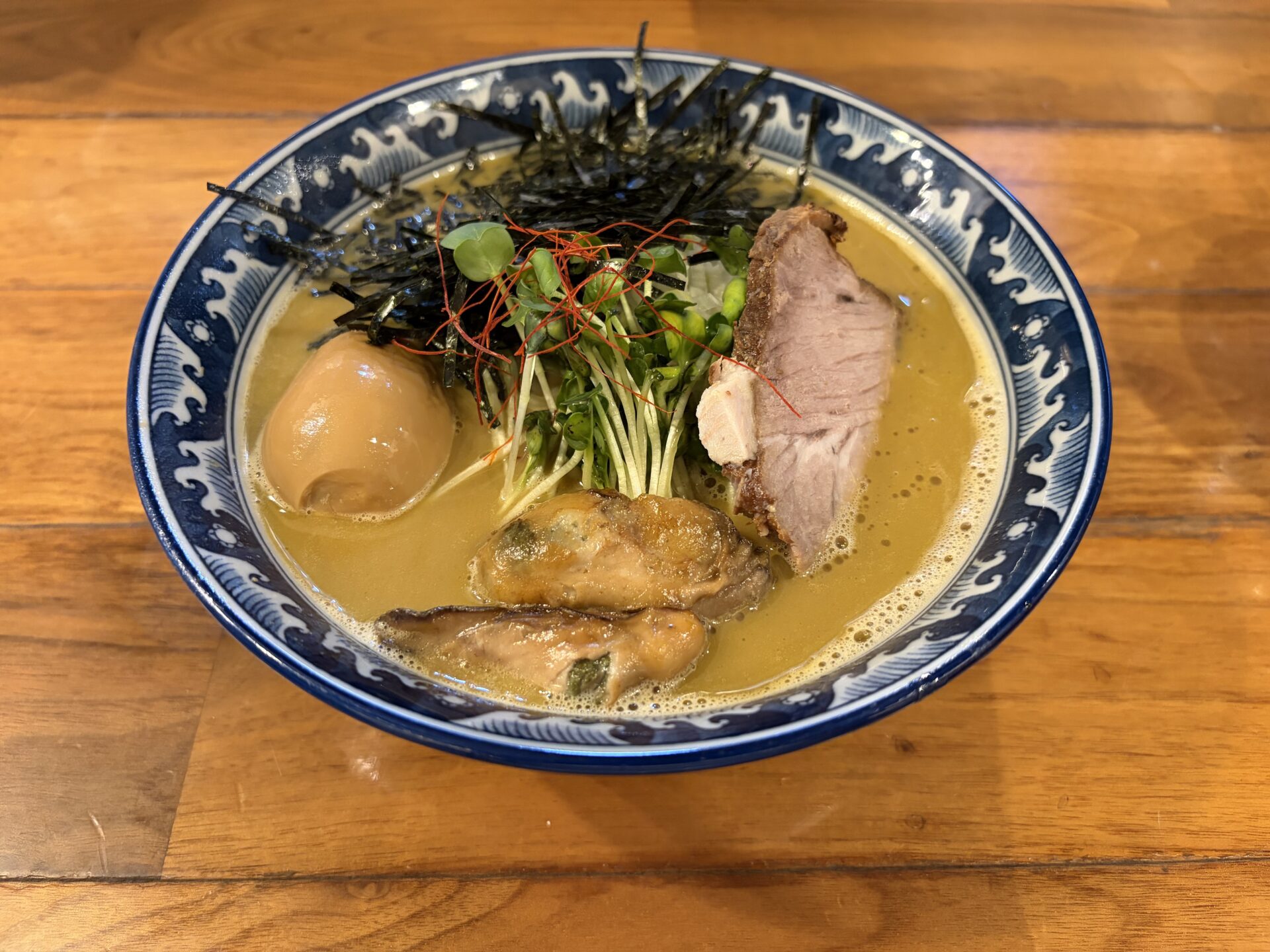 佐市麺の着丼。泡立つほど濃厚な黄土色のスープに、牡蠣・スープ色に染まった味玉・厚切りのチャーシュー・刻み海苔・かいわれ・糸唐辛子
