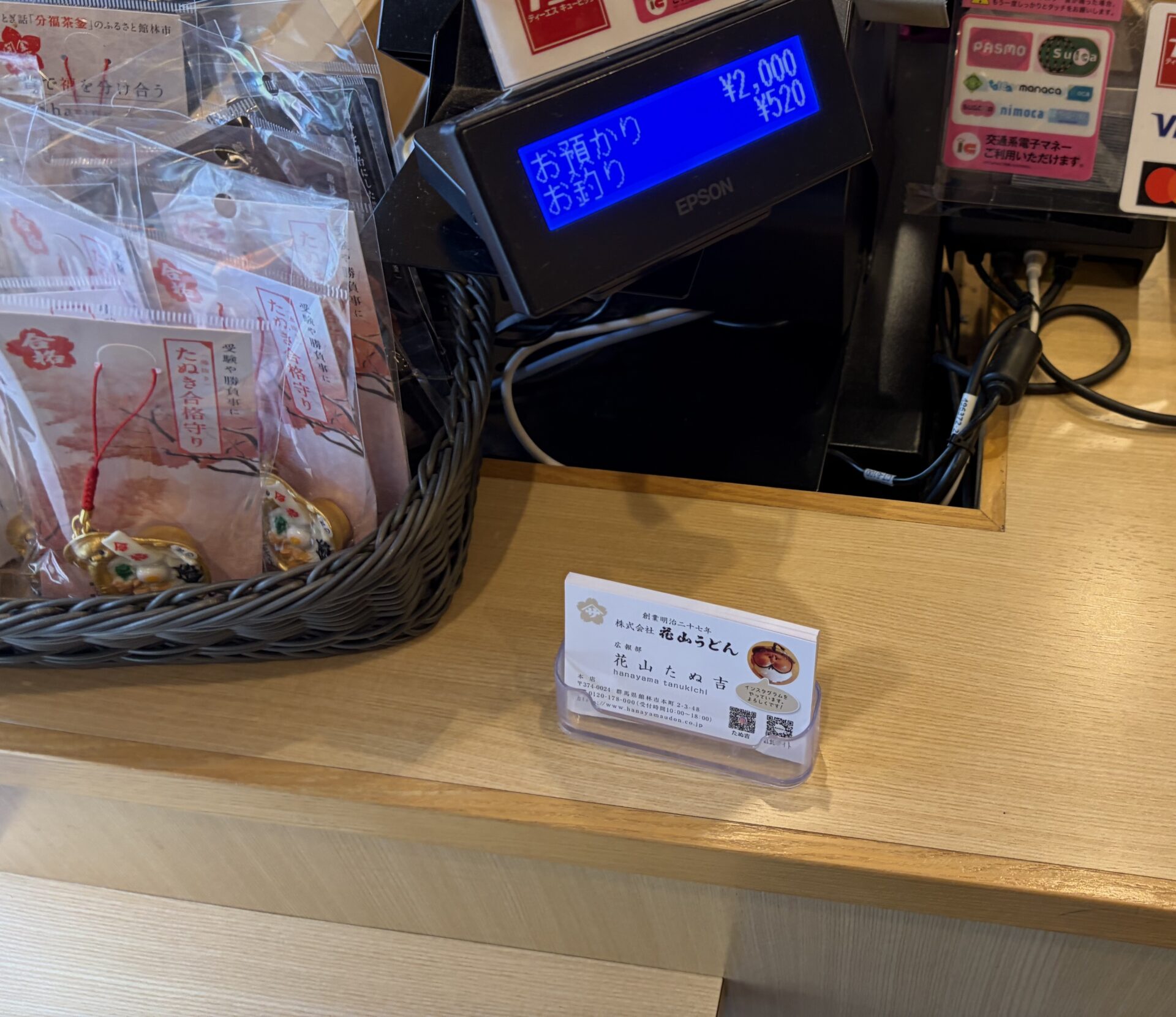 レジ。カード・交通系ICの掲示と、たぬきの合格守り