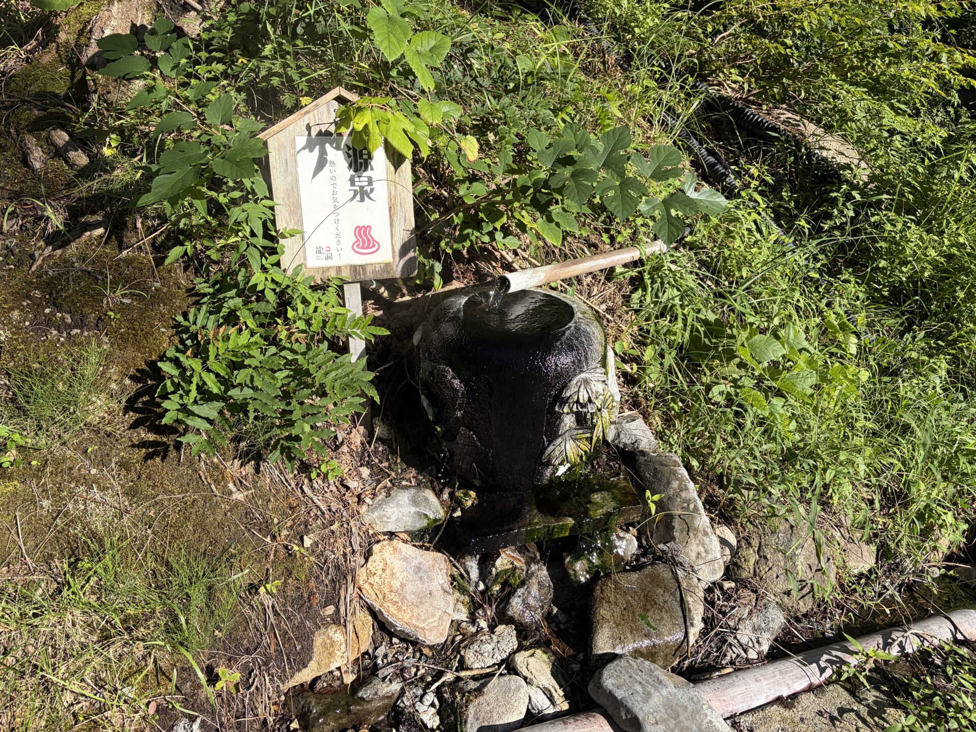 東館の裏手にある源泉の湧き口。「源泉 熱いのでお気をつけください!」の札と、黒い石の器に注ぐ源泉