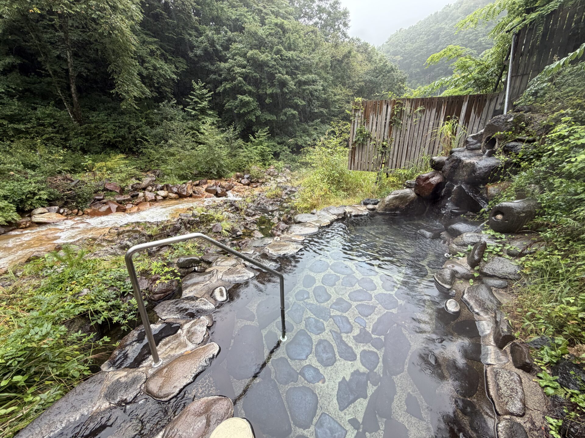 野天風呂の全景。左手を濁った川が流れ、岩の浴槽に湯が張られている