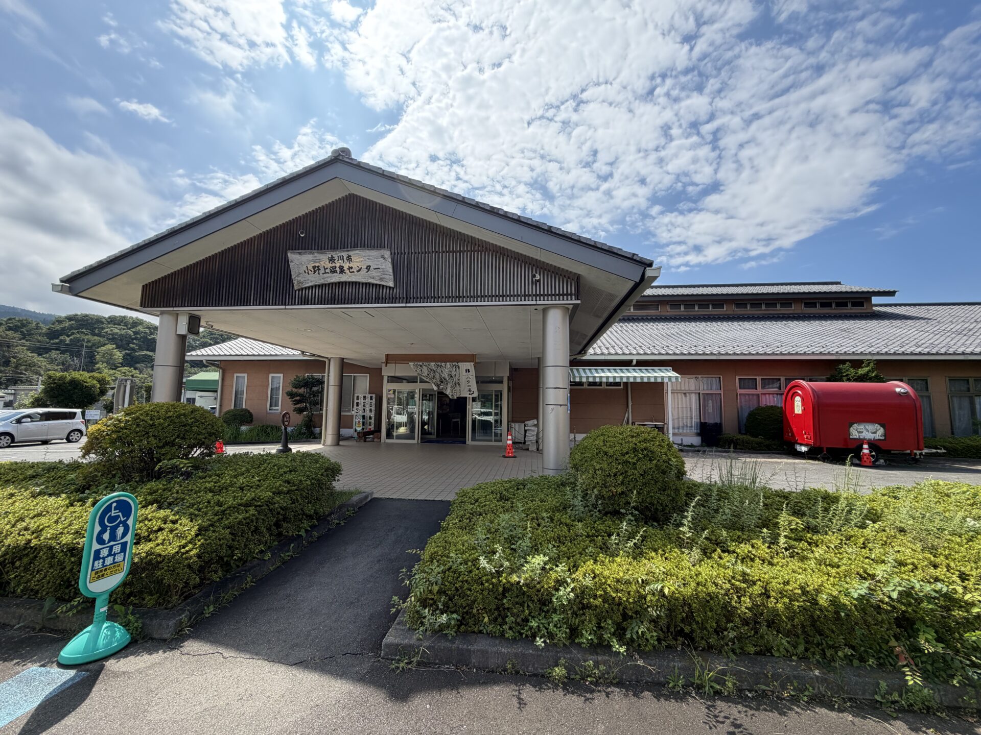 切妻の車寄せに木製看板「渋川市 小野上温泉センター」、入口に暖簾「ハタの湯」。JR吾妻線 小野上温泉駅前の外観