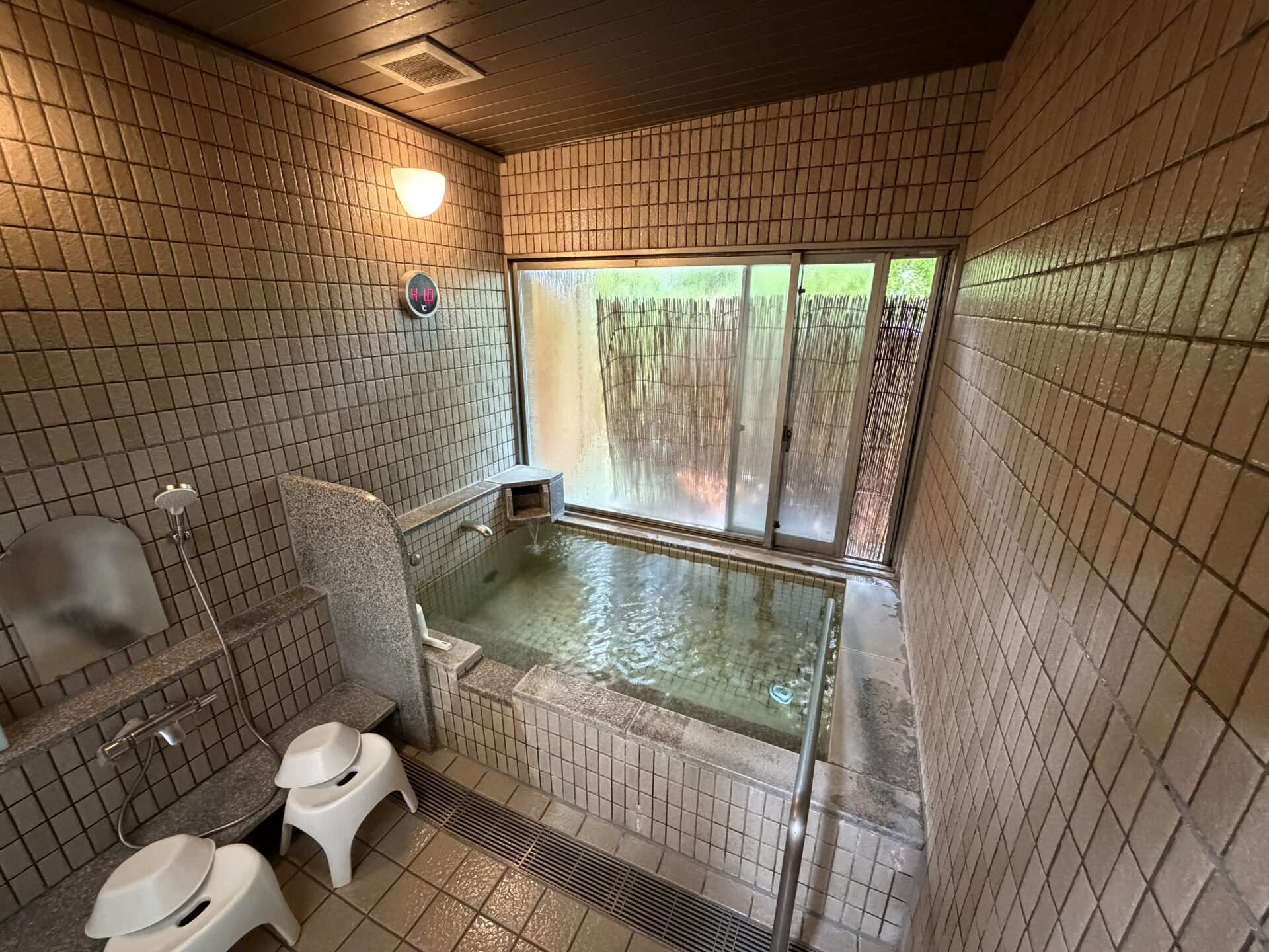 岩井堂の浴室。石縁の浴槽、洗い場1つ、簾の掛かった窓、壁の丸型デジタル湯温計