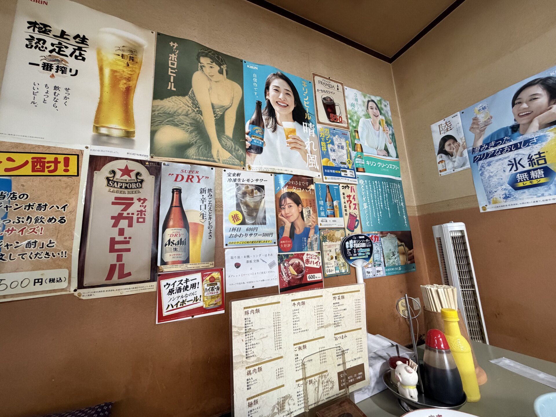 店内の壁。ビールやサワーのポスターの中ほどに「取り皿・お椀・トング・はさみ 鉄板交換」はタブレット【サービス】から、という貼り紙