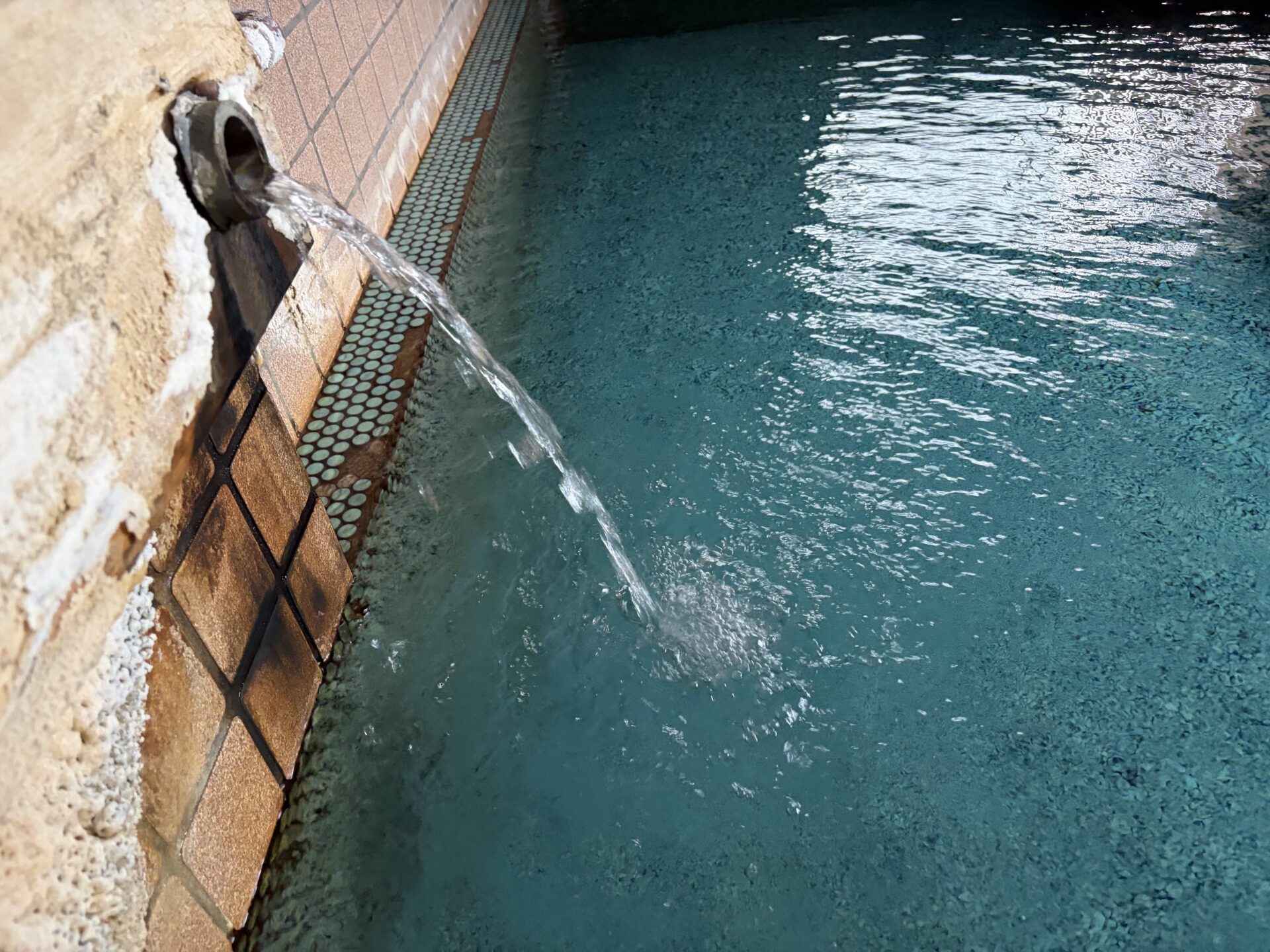 木枠の湯口を横から。鉄管の口に白い析出物が付き、湯が糸を引いて浴槽に落ちる