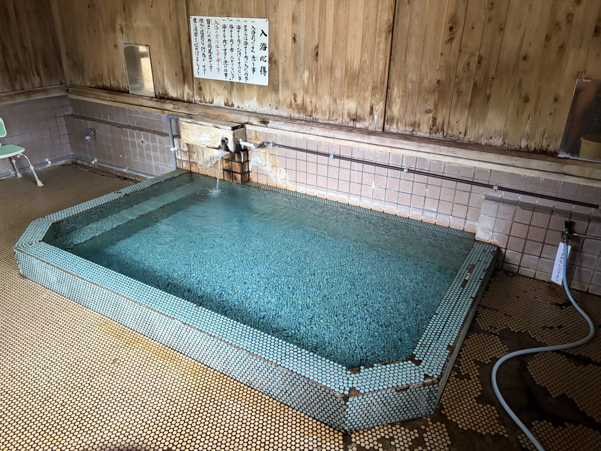 緑の丸タイルで縁取られた浴槽。奥の木枠の湯口から湯が落ち、右手の壁に水道の蛇口とホース