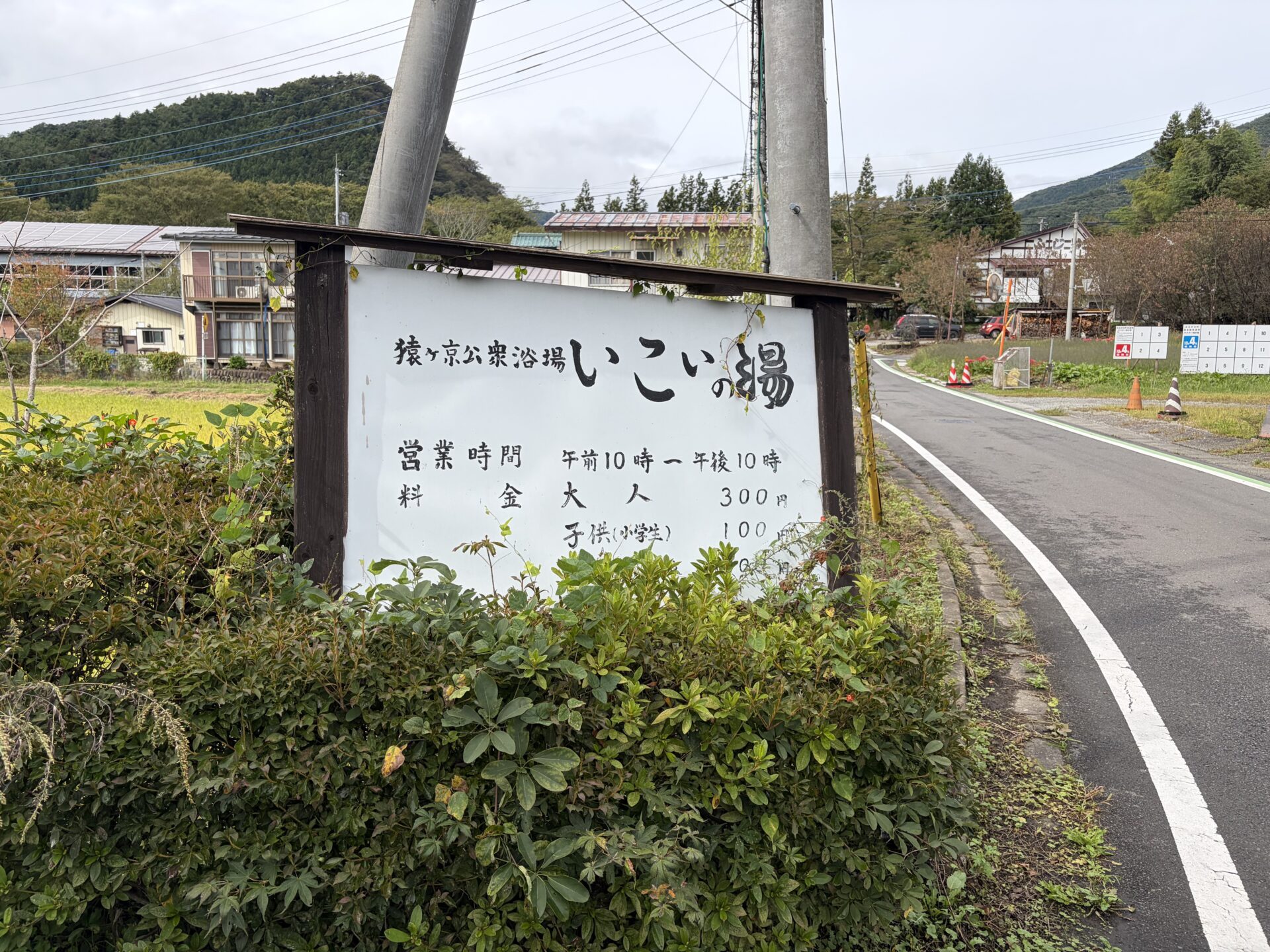 道路沿いの白い看板「猿ヶ京公衆浴場 いこいの湯」。営業時間と料金