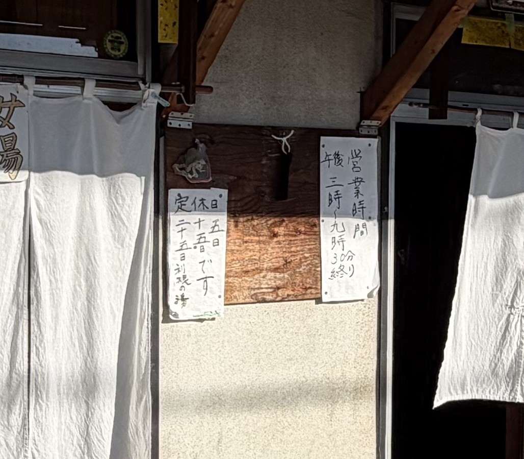 前橋の銭湯 利根の湯 男湯と女湯の暖簾の間の板壁に貼られた手書きの貼り紙。「定休日 五日 十五日 二十五日です 利根の湯」「営業時間 午後三時〜九時30分終り」
