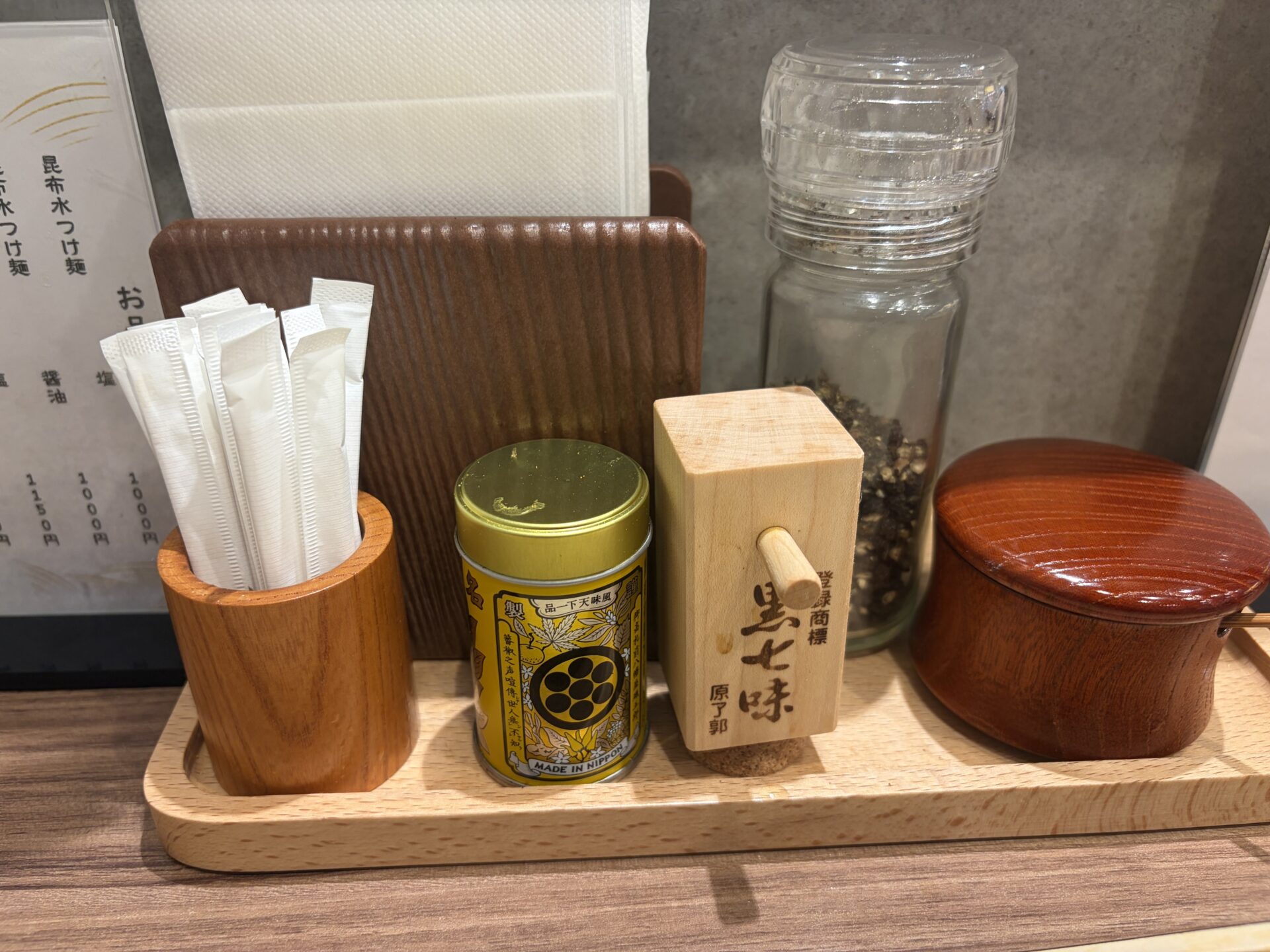 卓上の調味料。黄色い缶、「黒七味 原了郭」の木箱、ガラス瓶、藻塩の入った朱塗りの容器