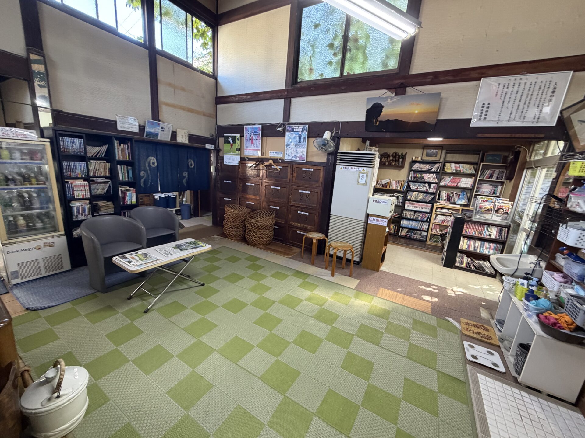 脱衣所。高窓のある高い天井、番号付きの木のロッカー、積まれた脱衣籠、漫画の棚