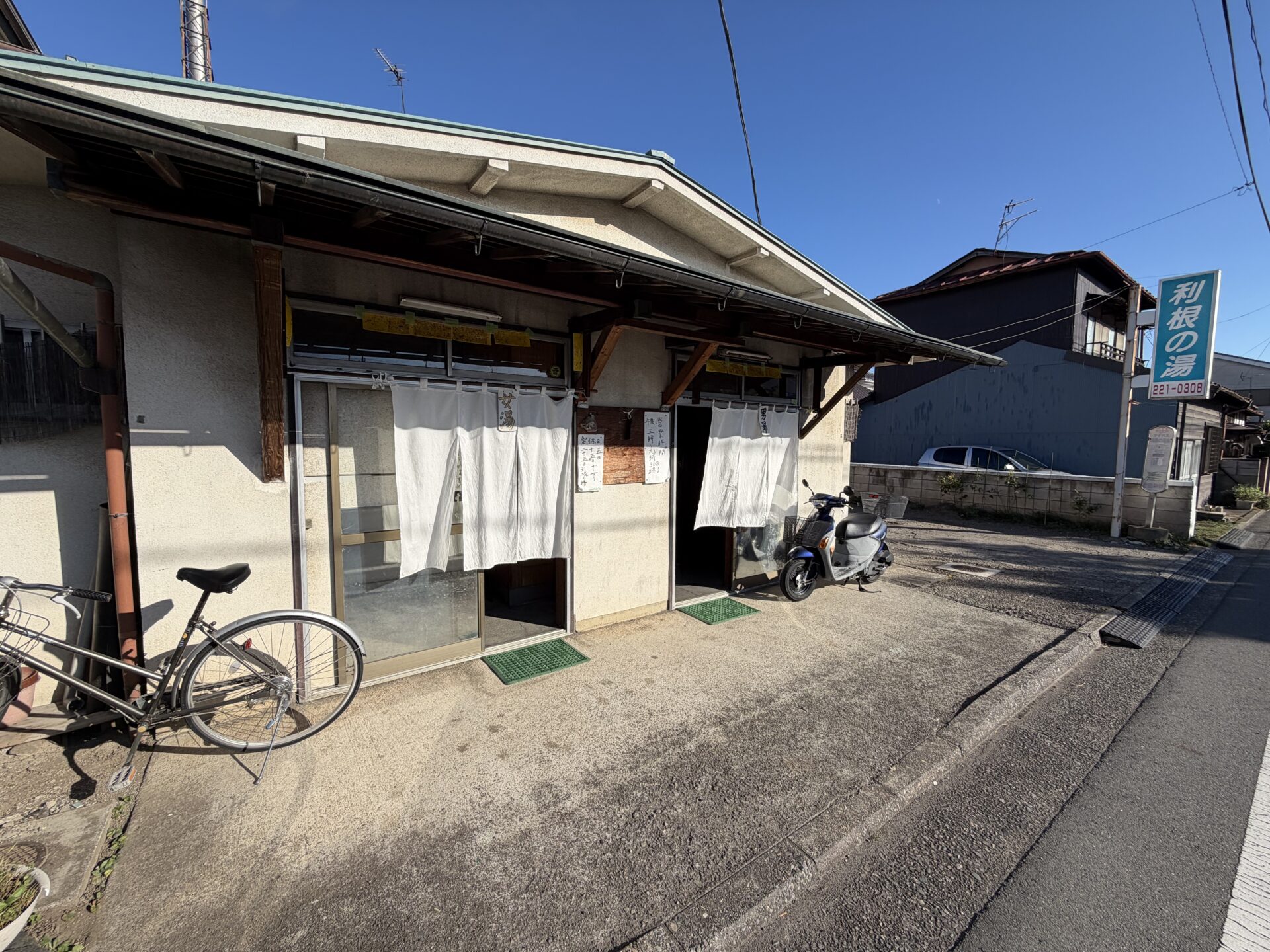前橋の銭湯 利根の湯 利根の湯の正面。建物に屋号は無く、白い暖簾2枚と手書きの貼り紙、道路脇の青い看板だけが目印